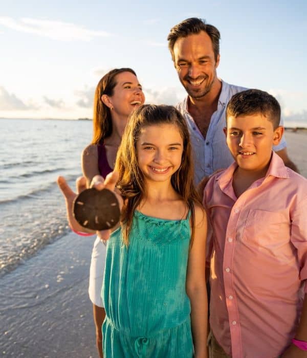 Four people stand on a beach at sunset. A girl in front holds up a sand dollar while the others smile in the background.