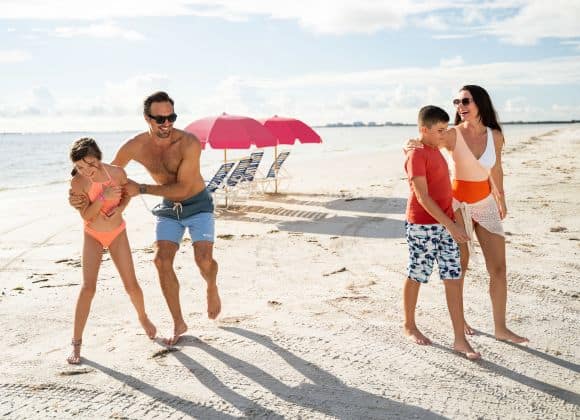 A family of four, dressed in swimwear, plays and laughs together on a sandy beach with two pink umbrellas and lounge chairs in the background.