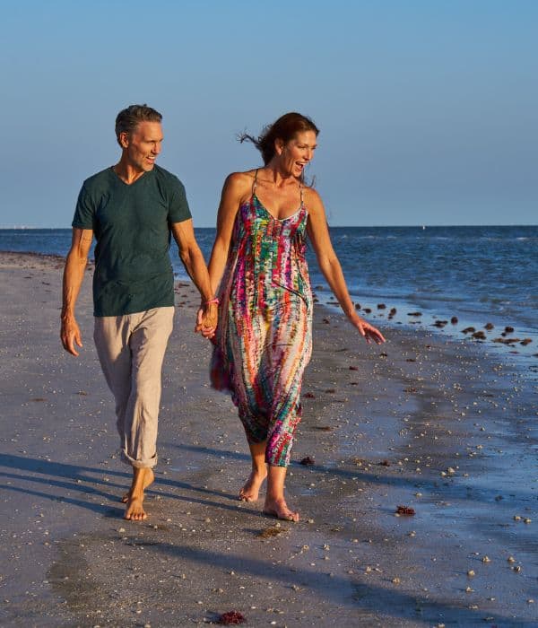 A man and woman walk barefoot, holding hands along a sandy beach near the ocean during daylight.