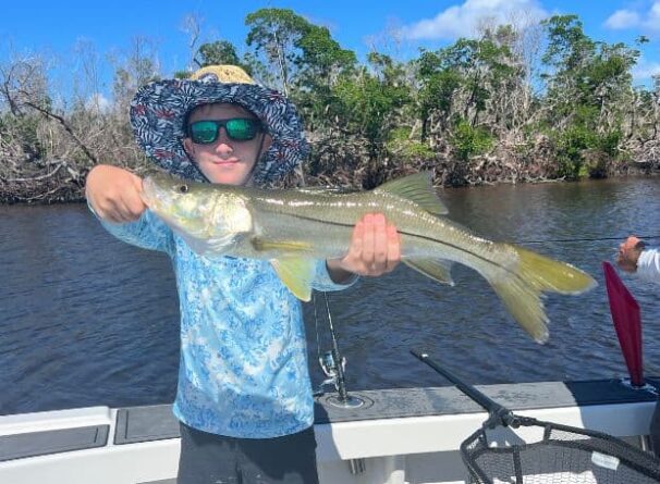 A boy wearing sunglasses and a hat holds a large fish on a boat, with trees and water visible in the background.