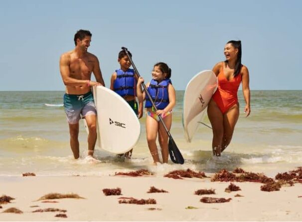 Four people, two adults and two children, walk out of the ocean carrying paddleboards and paddles on a sunny beach. The children wear life vests and everyone appears to be smiling.