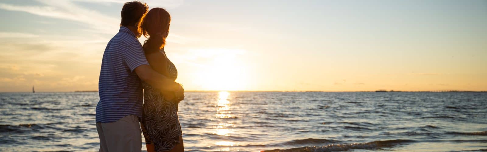 A couple stands on the beach, embracing as they look out at the ocean during a sunset.