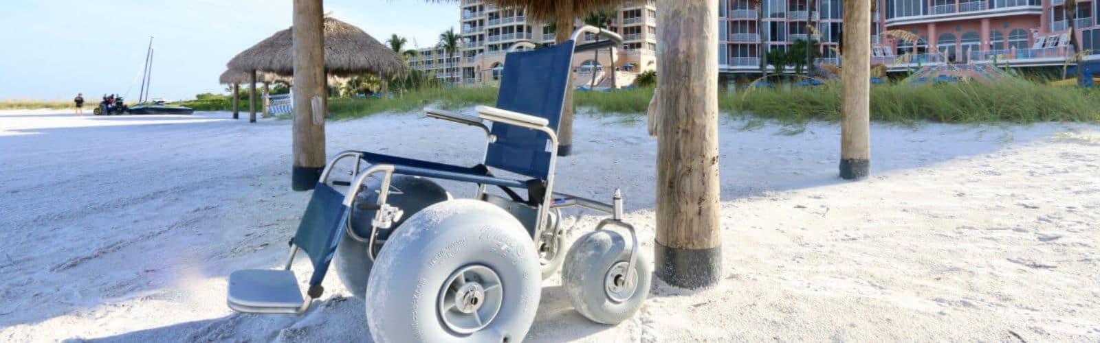 A beach wheelchair with large wheels is parked on white sand under a thatched umbrella near a resort building.