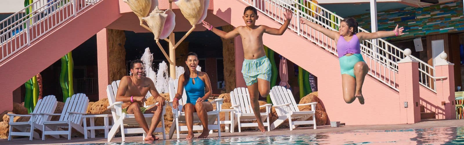 Two children jump into a swimming pool while two adults sit on white chairs nearby at a colorful outdoor pool area.