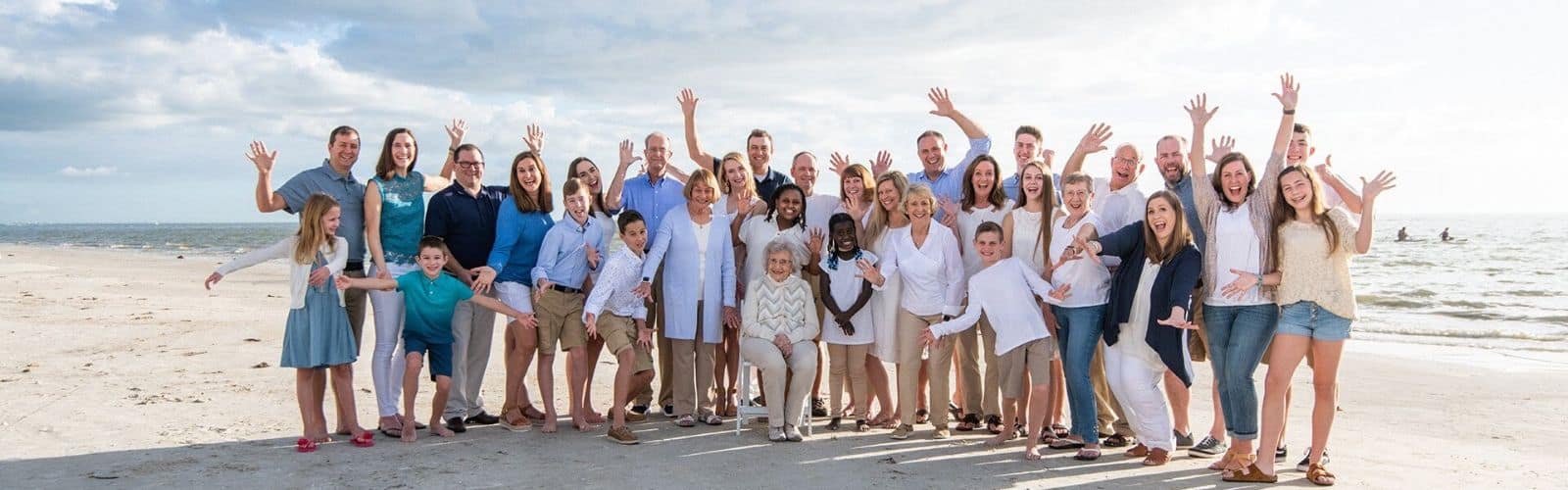 A large group of people posing and smiling with arms raised on a sandy beach near the ocean, under a partly cloudy sky.