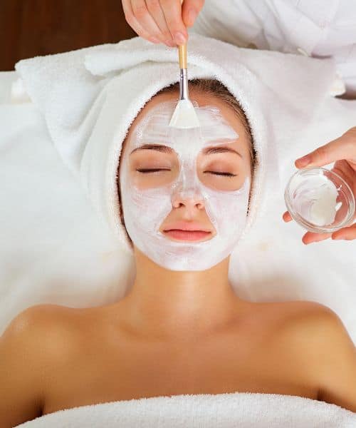 Person lying down with a towel wrapped around their head while a facial mask is being applied to their face with a brush.