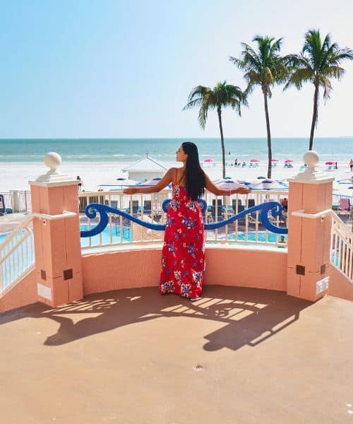 A woman in a red floral dress stands on a balcony overlooking a pool, beach, and palm trees on a sunny day.