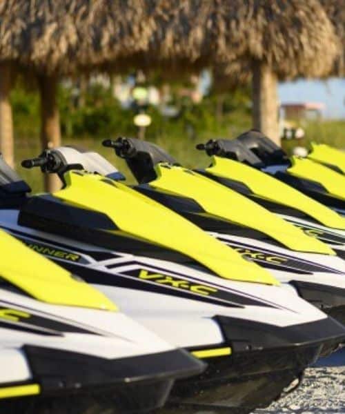 A row of yellow and white jet skis are lined up on a sandy beach with thatched huts and greenery in the background.