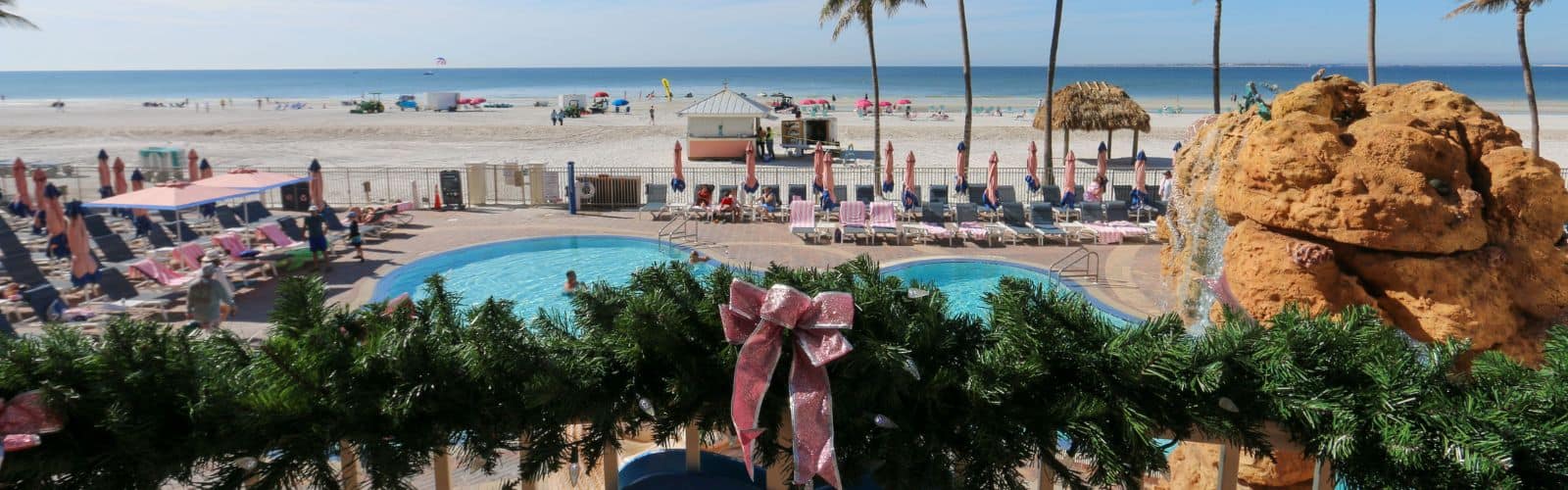 View of a beachfront resort with pools, lounge chairs, umbrellas, and holiday garland with pink bows in the foreground; beach and ocean in the background.
