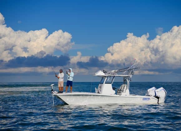 Two people are fishing on a white motorboat in calm open water under a partly cloudy blue sky.