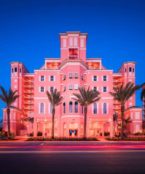 A pink, multi-story hotel building with palm trees in front, illuminated at dusk, with blurred car lights on the street in the foreground.