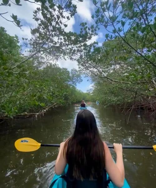 A person with long dark hair kayaks through a narrow waterway surrounded by dense green trees, following others in separate kayaks under a partly cloudy sky.