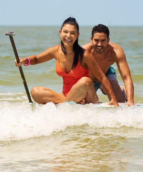 A woman in an orange swimsuit and a man in swim trunks paddle together on a board in the ocean, both smiling and enjoying the water.