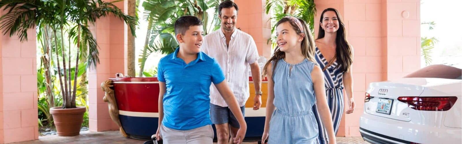 A family of four, two adults and two children, walk together near a white car outside a building with tropical plants and a red boat decoration.