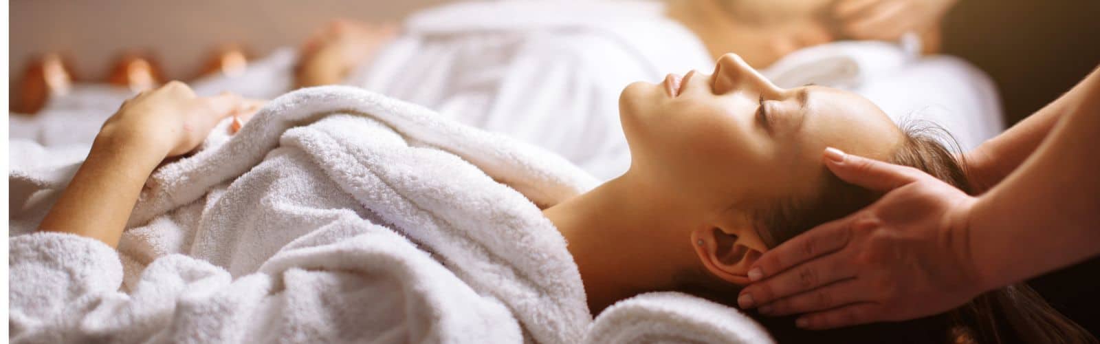 A person in a white robe lies on a massage table receiving a head massage, with another person blurred in the background.