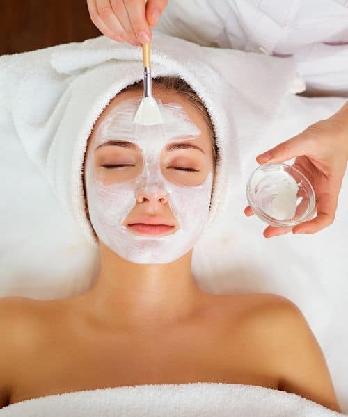 Person lying down with eyes closed, wearing a towel on their head, while a facial mask is being applied with a brush.