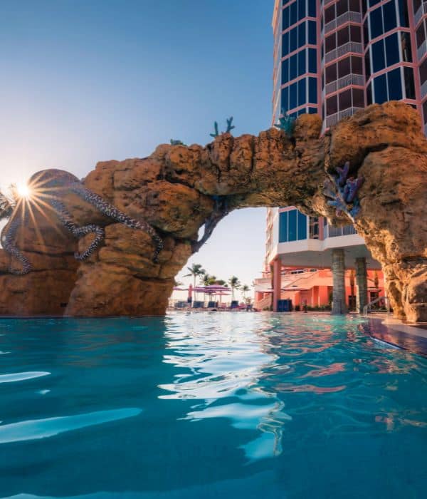 A pool with a rock archway decorated with sea creatures, adjacent to a tall modern building, under a clear sky at sunset.