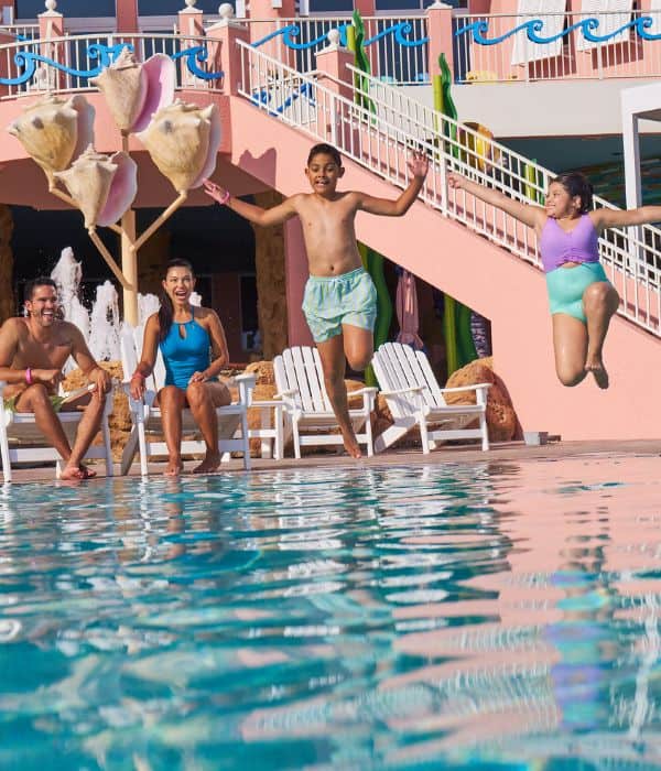 Four people sit by a pool while two children jump into the water. The background includes pink walls, white chairs, and a cotton candy decoration.