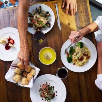 Two people dining at a wooden table with plates of pasta, seafood, salad, bread, dessert, olive oil dip, and glasses of wine.