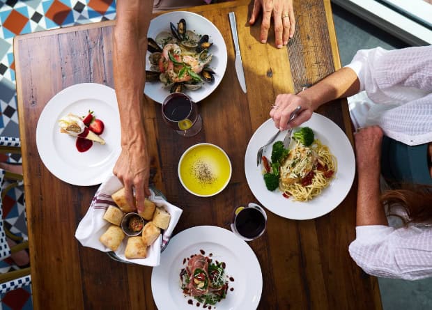 Two people dining at a wooden table with plates of pasta, seafood, salad, bread, dessert, olive oil dip, and glasses of wine.