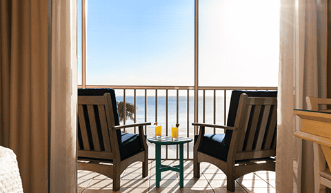 Two wooden chairs with blue cushions face a small table holding drinks on a balcony overlooking a calm ocean under a clear, sunny sky.