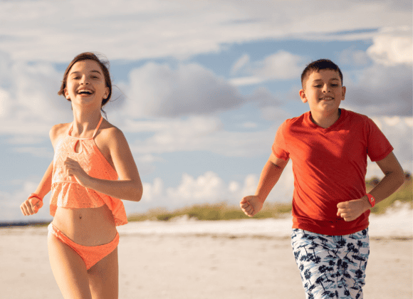 Two children, a girl in a peach swimsuit and a boy in a red shirt with patterned shorts, run on a sandy beach under a partly cloudy sky.