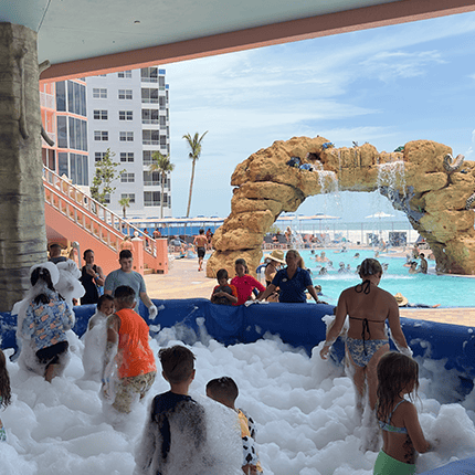 Children and adults play in a foam-filled area near an outdoor pool, with a rocky waterfall feature and palm trees in the background.