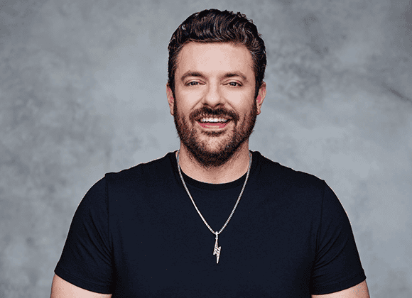 A man with dark hair and a beard wears a black t-shirt and a silver necklace, smiling in front of a gray background.