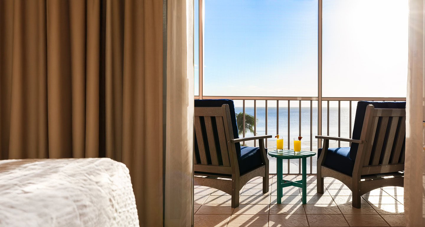 Two wooden chairs and a small table with drinks on a balcony overlook a sunny ocean view, seen from inside a room with beige curtains and bedding.
