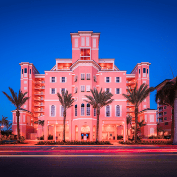 A pink multi-story hotel with palm trees in front, illuminated at dusk, with light trails from passing cars on the street.