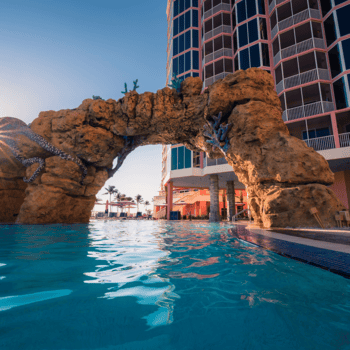 A swimming pool with a rock arch featuring sculptures, next to a multi-story hotel building, under a clear blue sky.