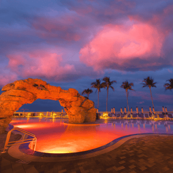 A resort swimming pool with rock formations and palm trees, illuminated by pink and orange lights, under a dramatic sunset sky with clouds.