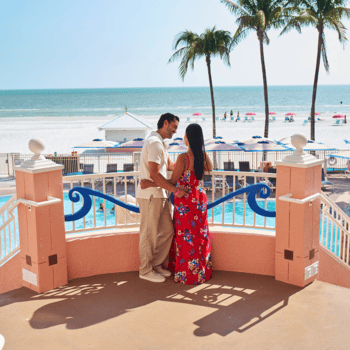 A couple stands on a terrace overlooking a pool, white sand beach, and palm trees with people and umbrellas in the background.