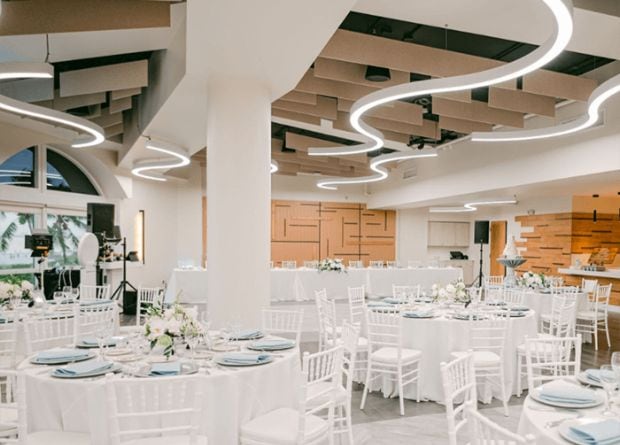 Modern banquet hall with white chairs and tables set for an event, featuring contemporary ceiling lights and neutral decor.