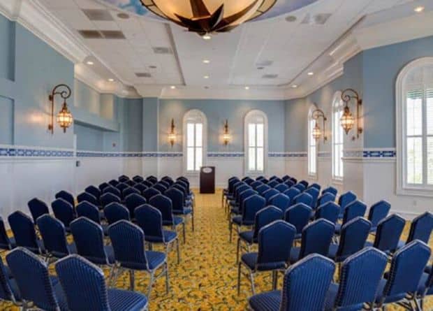 Rows of blue chairs are arranged facing a podium in a bright, empty conference room with blue walls, arched windows, and decorative wall sconces.