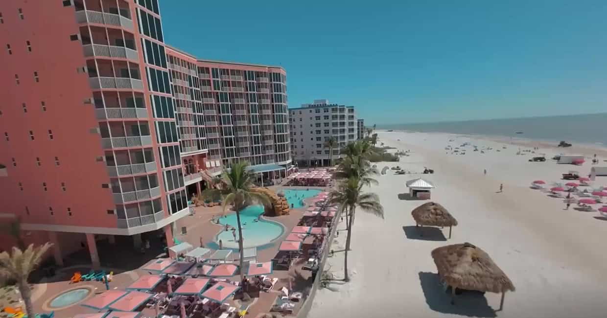 A beachfront resort with a pink multi-story building, outdoor pool area, palm trees, cabanas, and beach chairs along a sandy shoreline under a clear blue sky.