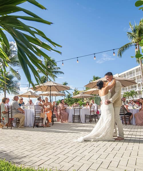A bride and groom dance outdoors on a sunny day, surrounded by seated guests at round tables under umbrellas and string lights, with palm trees and a hotel in the background.