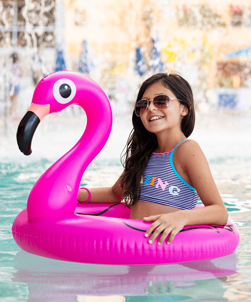 A young girl wearing sunglasses sits in a pool inside a pink flamingo float. She is smiling and wearing a striped swimsuit. The background shows water and blurred poolside details.