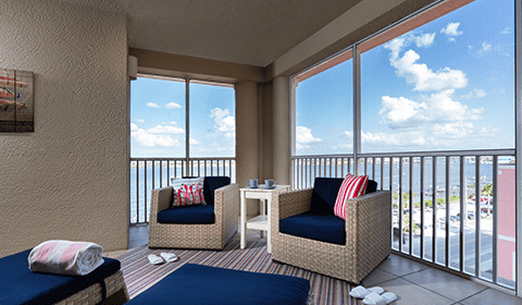 A furnished balcony with wicker chairs, a small table, and folded towels overlooks a body of water and a cityscape under a partly cloudy sky.
