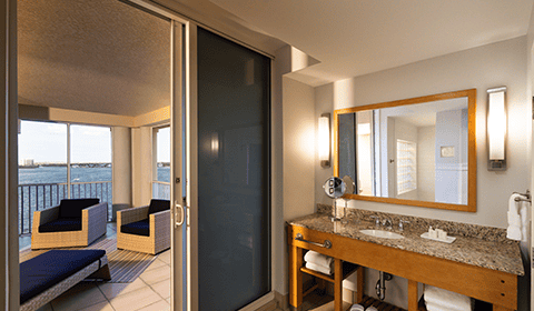 Hotel bathroom with a large mirror and sink, next to a sliding glass door leading to a balcony with chairs overlooking a body of water.