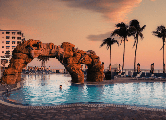 Outdoor swimming pool with a rock arch, palm trees, and lounge chairs at sunset, with one person in the water and others standing nearby.