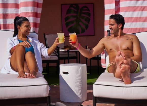 Two people relax on lounge chairs by a small table, smiling and toasting with drinks in hand; striped cabana curtains and a leaf painting are in the background.