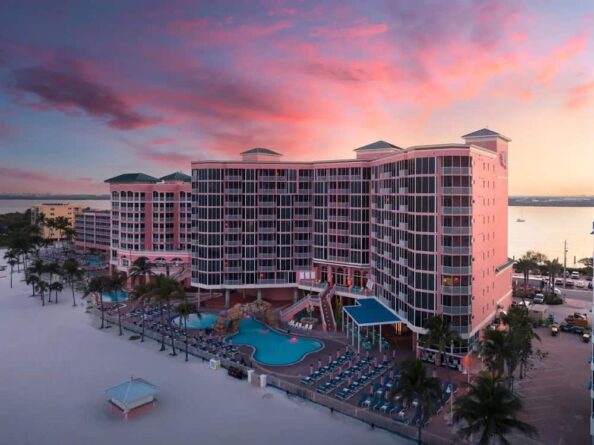 A large pink beachfront resort with multiple stories, a central swimming pool, palm trees, and lounge chairs, seen at sunset with a colorful sky in the background.