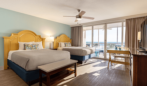 Hotel room with two queen beds, a ceiling fan, a TV, a desk, and a balcony with an ocean view through large sliding glass doors.