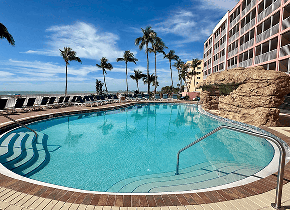 Outdoor swimming pool with clear blue water, surrounded by lounge chairs, palm trees, and a multi-story hotel building under a sunny sky.