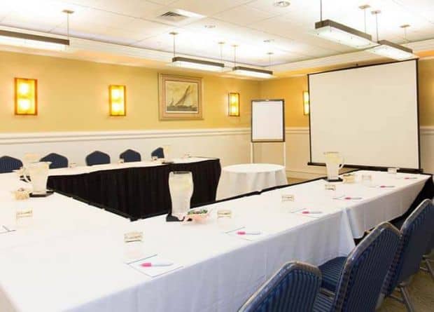 Conference room with U-shaped table setup, chairs, water pitchers, name cards, a projection screen, whiteboard, and wall lights in a well-lit space.