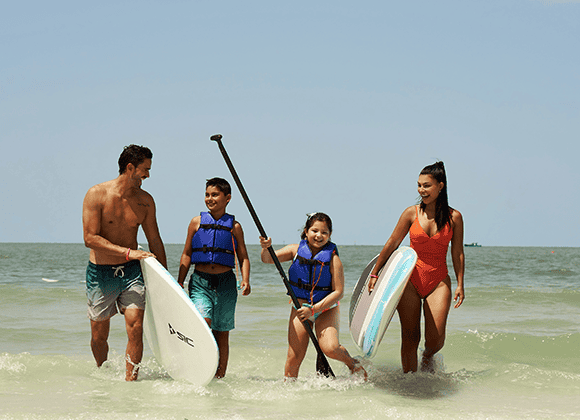 A man, woman, and two children in life vests walk through shallow ocean water carrying paddleboards on a sunny day.