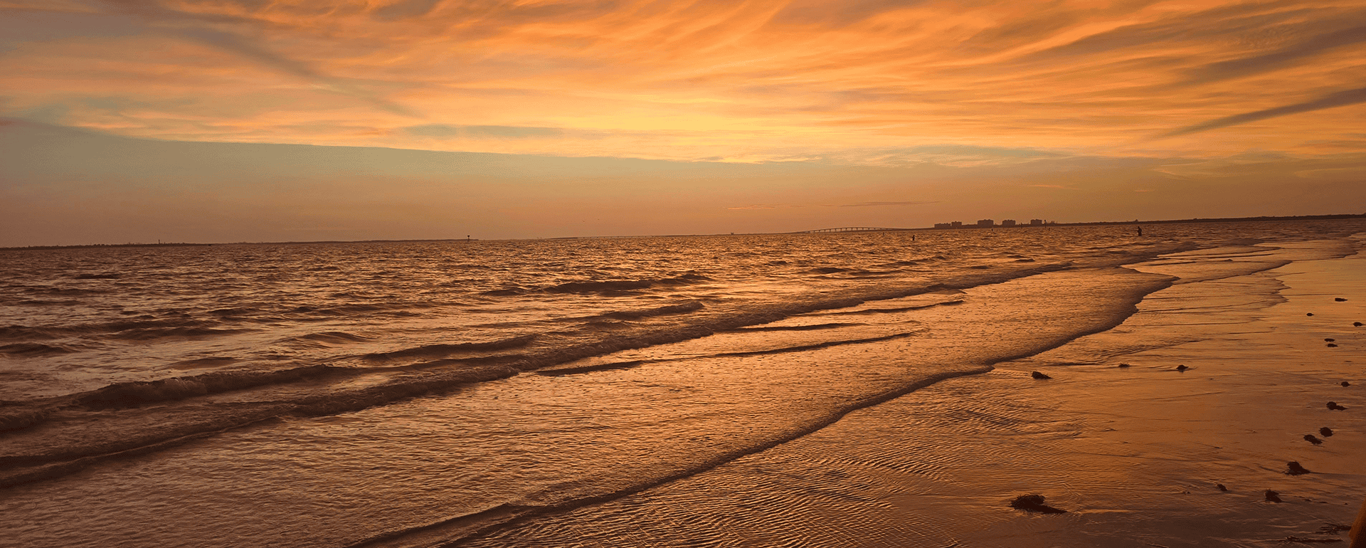 A sandy beach at sunset with orange clouds, gentle waves, and distant buildings on the horizon.