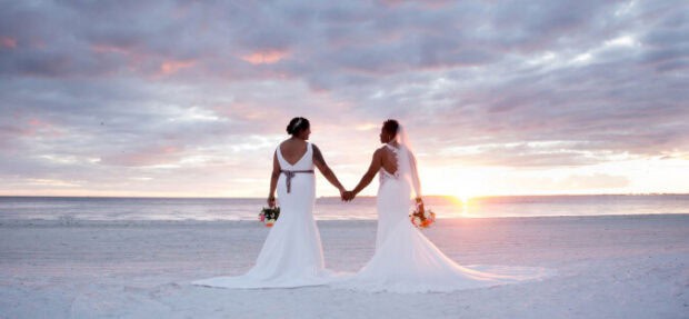 Two women in wedding dresses hold hands and stand on a beach at sunset, each holding a bouquet, with the ocean and cloudy sky in the background.