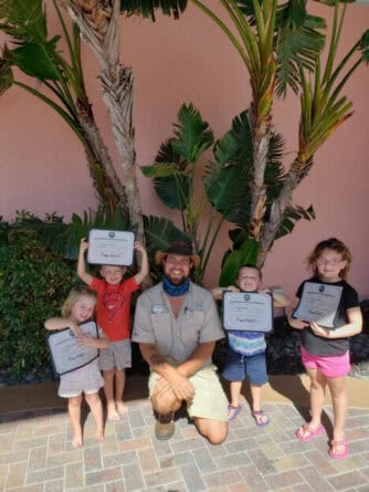 An adult kneels in front of palm trees with four children standing, each holding a certificate and smiling at the camera.